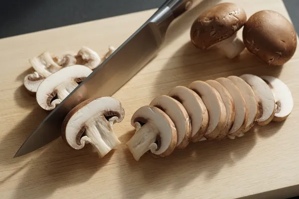 Hands slicing cremini mushrooms into even quarter-inch slices on a wooden board