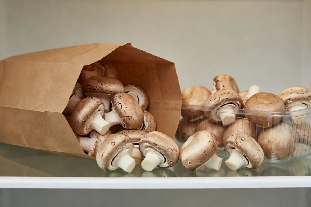 Cremini mushrooms in an open paper bag beside a vented carton on a refrigerator shelf